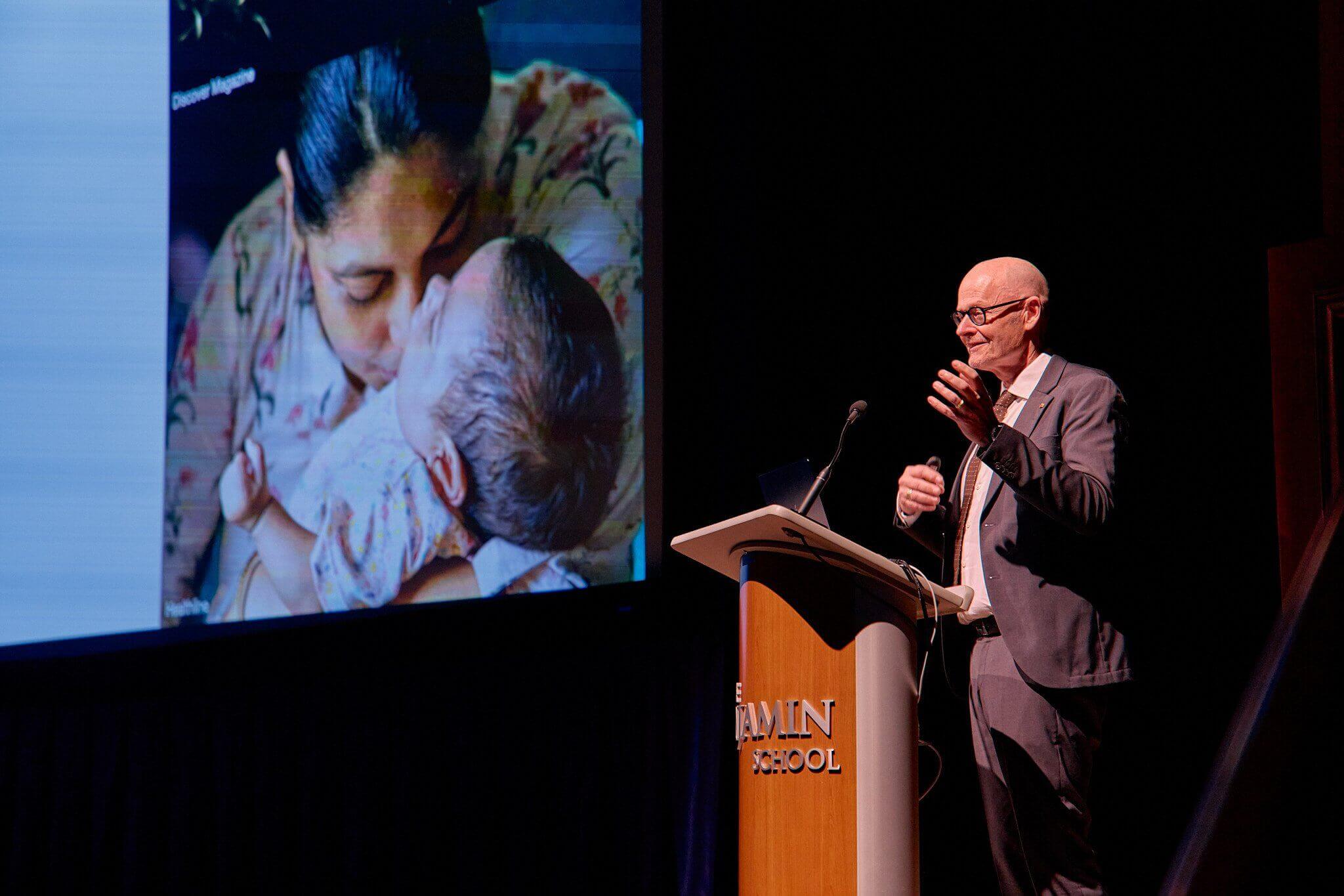 DSC09477-1 Prof. Dr. Hansson speaks behind a podium. An image of a woman and baby are projected on the screen to his right.